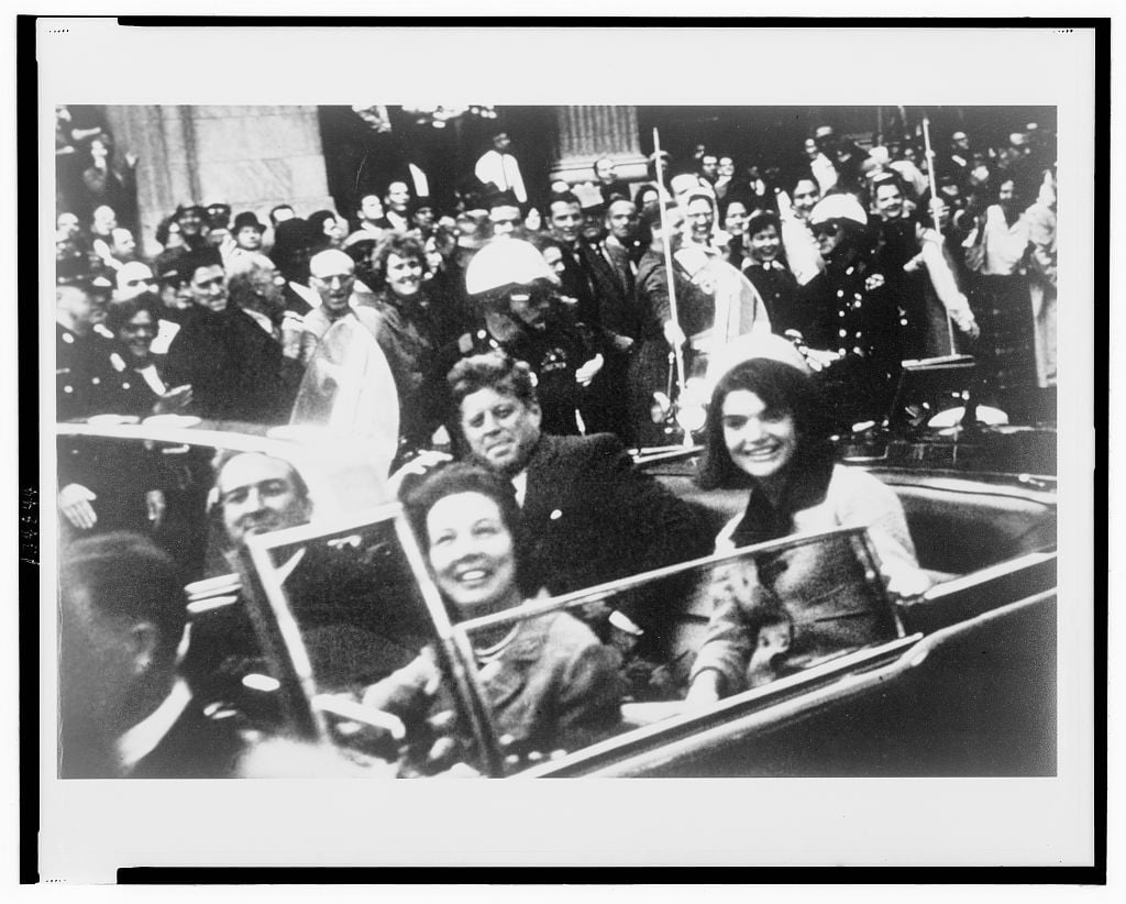 Black/white photo of four well-dressed people in black convertible with crowd cheering in background